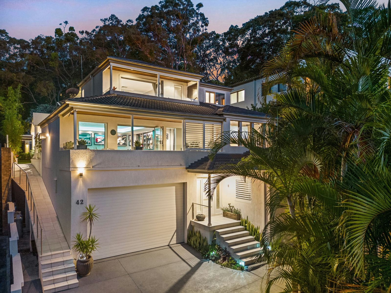 Modern three-story home at dusk with every window lit, framed by palms
