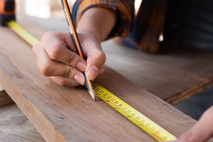 Carpenter's hand marking a board with a pencil against a tape measure