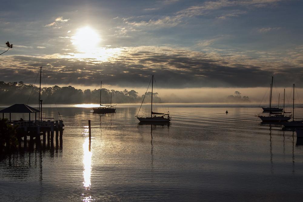 Fog lifting off a calm harbor at sunrise with boats at anchor
